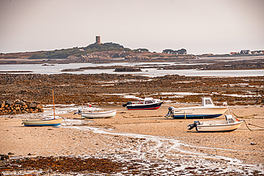 A scenic coastal view at low tide on Guernsey in the Channel Islands, showing small boats resting on the sandy shore with Fort Saumarez in the distance