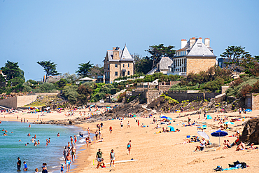 A lively summer scene on the sandy Plage du Mihinic, Saint Mao, Brittany, France, where sunbathers and swimmers enjoy the warm weather. In the background, elegant Belle Époque villas
