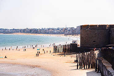 People walking along the sandy beach below the fortified walls of Saint-Malo, Brittany, France, with wooden breakwaters and the walled city in the background