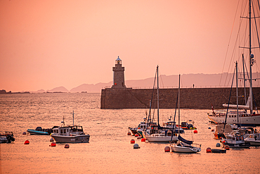 : Golden hour at Saint Peter Port Guernsey with yachts anchored in the harbour lighthouse on the breakwater and pastel colored sunset sky across the Channel Islands coastline