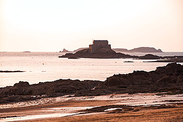 Silhouette of Grand Bé tidal island and ancient fort beyond Saint-Malo’s rocky beach with pastel sunset sky reflecting Brittany’s maritime and literary allure