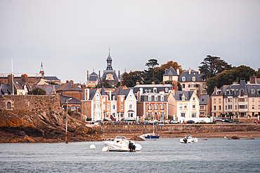 Historic stone houses and waterfront buildings line the riverbank of the Rance estuary .Le Rosais, Brittany, France. Yachts and small boats are anchored along the calm shoreline.