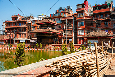 Plant pots and brick buildings surrounding the lake, and bamboo logs stored at the vibrant green Bholachhe Pond in the core of Bhaktapur, Kathmandu Valley, Nepal, Asia