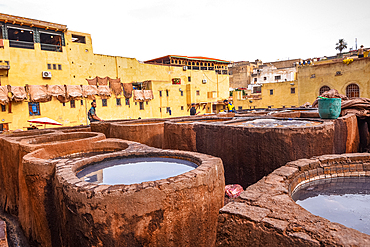 Tannery workers overseeing dye pits in a traditional leather tannery in Fes, Morocco, with yellow walls in the background.