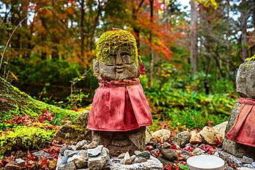 A moss-covered Warabe-Jizo statue wearing a red bib, surrounded by vibrant autumn leaves and peaceful greenery.