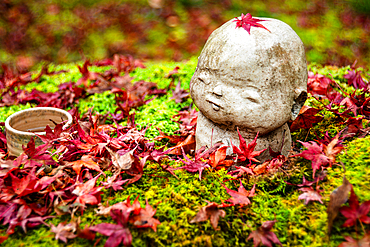 A tranquil Warabe-Jizo statue resting on mossy ground, topped with a red maple leaf and surrounded by autumn foliage.