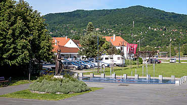 Visegrád, Hungary – View of a quiet town park with benches, a statue, and red-roofed houses framed by forested hills in the Danube Bend. A peaceful civic space blending nature, memory, and local charm.