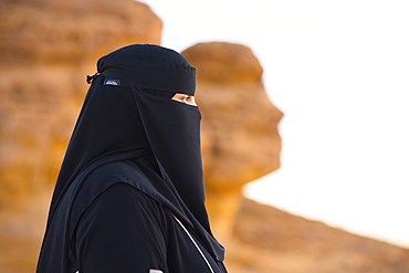 Woman wearing a Niqab posing in front of the 'Face Rock', a sandstone rock formation shaped like the human head profil, within the UNESCO World Heritage Site of Hegra, AlUla, Medina Province, Saudi Arabia, Middle East