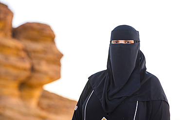 Woman wearing a Niqab posing in front of the 'Face Rock', a sandstone rock formation shaped like the human head profil, within the UNESCO World Heritage Site of Hegra, AlUla, Medina Province, Saudi Arabia, Middle East