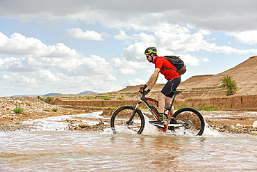 Rider on mountain bike with electric assistance crossing a river with ford on a track between Ouarzazate and Ait Ben Haddou, Ounila River valley, Ouarzazate Province, region of Draa-Tafilalet, Morocco, North West Africa