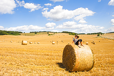 Young girl sitting on a straw roll in an harvested field, Allier department, Auvergne-Rhone-Alpes regionFrance, Europe