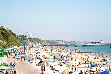 Crowded beach on a sunny day with people sunbathing and swimming, colorful umbrellas, and a pier in the distance in Bournemouth, Dorset, England, United Kingdom, Europe