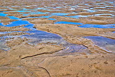 A close-up view of a sandy beach with shallow water pools. The sand features ripples and patterns created by the tide, with reflections of the sky visible in the water. The overall scene conveys a serene coastal atmosphere.