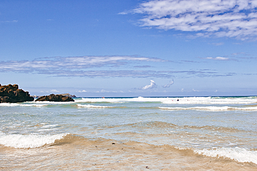 A serene beach scene featuring gentle waves lapping at the shore under a clear blue sky with scattered clouds. The water is calm, and a few surfers can be seen in the distance, enjoying the ocean.