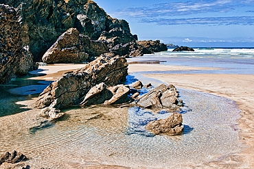 A serene beach scene featuring large rocky formations along the shore. The tide is low, revealing sandy areas and shallow water pools. The sky is partly cloudy, and the ocean waves gently lap at the beach.