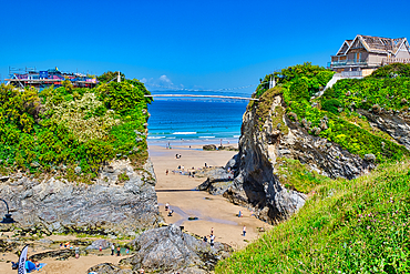 A scenic beach view featuring cliffs on either side, with people walking on the sandy shore. The ocean is calm and blue, and a bridge can be seen in the distance. Lush greenery adorns the cliffs, and a clear blue sky completes the picturesque scene.