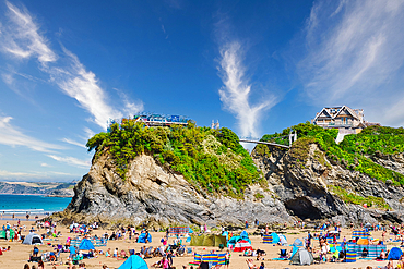 A vibrant beach scene featuring numerous people enjoying the sun, colorful beach tents, and rocky cliffs in the background. The sky is bright with wispy clouds, and there are houses perched on the cliffs overlooking the beach.