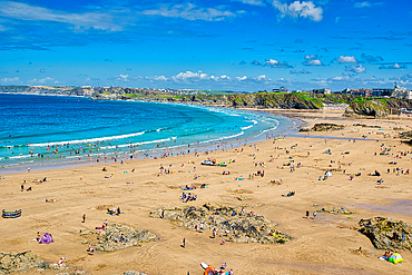 A vibrant beach scene with golden sand and clear blue waters. People are enjoying various activities like swimming, sunbathing, and playing on the beach. The coastline features cliffs and greenery in the background under a bright blue sky with fluffy clou