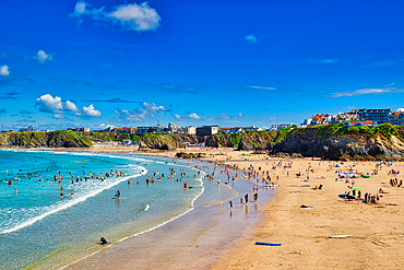 A vibrant beach scene with people enjoying the sun and water. The sandy shore curves along the coastline, with cliffs in the background and a clear blue sky dotted with clouds. Surfboards and beach umbrellas are visible, creating a lively atmosphere.