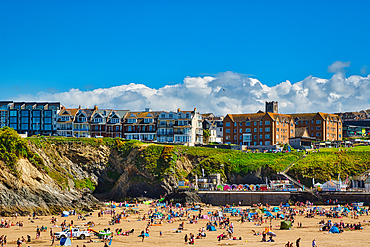 A vibrant beach scene with numerous people enjoying the sun on a sandy shore. Colorful beach umbrellas and towels are scattered across the sand. In the background, a row of buildings overlooks the beach, with a clear blue sky and fluffy clouds above.