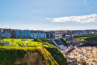 A vibrant coastal scene featuring a sandy beach filled with people enjoying the sun. In the background, there are colorful beach huts and a row of buildings overlooking the beach. The sky is clear with a few clouds, and lush green grass is visible in the