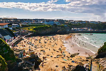 A vibrant beach scene with numerous people enjoying the sunny weather. The sandy shore is dotted with colorful umbrellas and beach towels, while waves gently lap at the coastline. In the background, there are houses and buildings on a hillside, under a pa