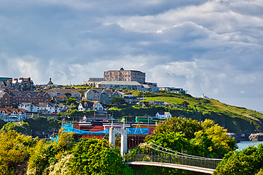 A scenic view of a coastal town with a hotel on a hill, surrounded by lush greenery and colorful boats in the harbor. The sky is partly cloudy, creating a dramatic backdrop.