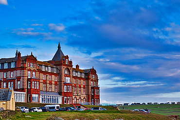 A historic red brick hotel building with a turret, set against a blue sky with scattered clouds. The hotel is situated on a grassy hill, with several parked cars in the foreground and a distant view of the coastline.