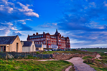 A scenic view of a large, historic hotel building with red brick architecture, set against a backdrop of blue skies and clouds. In the foreground, there are smaller, simpler buildings and a pathway leading towards the hotel. The landscape is grassy and op