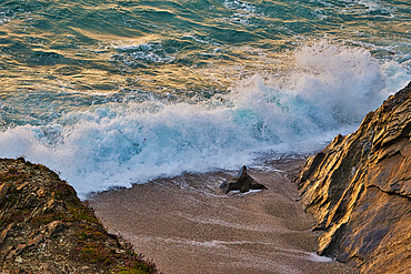 A scenic view of ocean waves crashing against rocky cliffs at sunset. The water is a mix of turquoise and white foam, creating a dynamic contrast with the sandy beach and rocky shoreline. The golden light of the setting sun enhances the natural beauty of