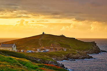 A scenic coastal landscape featuring a hill with a lighthouse at the top, surrounded by the ocean. The sky is painted with warm hues of orange and yellow during sunset, casting a beautiful glow over the scene. In the foreground, there are grassy areas and