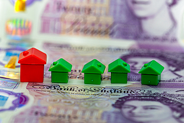 Five miniature house models, one red and four green, are arranged on top of a stack of British pound banknotes. The banknotes are out of focus, suggesting the concept of cost or value.