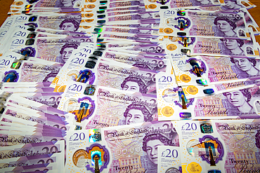 A large spread of British twenty-pound banknotes arranged on a flat surface. The notes feature intricate designs, holographic elements, and the portrait of Queen Elizabeth II. The colors are predominantly purple with gold accents.