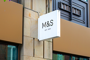 A close-up of a modern retail store sign displaying the letters 'M&S' with the text 'EST. 1884'. The sign is mounted on a stone wall, showcasing a contemporary design. The background features a brown panel, indicating a stylish storefront.