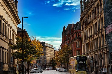 A city street with tall brick buildings, a yellow double-decker bus, and cars parked on the side of the road. The sky is blue with white clouds in Manchester, UK.