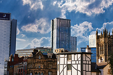 A city skyline with a mix of modern and historic buildings, including a tall glass skyscraper, a church, and a timber-framed building. The sky is cloudy, with some sun shining through in Manchester, UK.