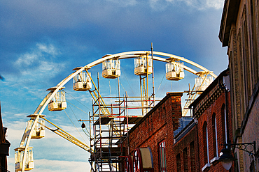 A partially constructed Ferris wheel is positioned in the background, with scaffolding visible and several empty cabins. In the foreground, two brick buildings flank the wheel, creating a frame. The sky is a hazy blue with white clouds in York, North Yor