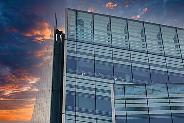 A modern glass and steel building against a colorful sunset sky. The building's facade features large windows and a grid pattern, while the sky is a blend of orange, purple, and blue.