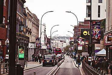 A street in London, UK, lined with shops and buildings, with a red double-decker bus driving down the center lane. People walk along the sidewalk, and streetlights curve over the street.