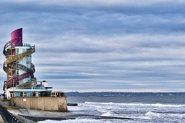 Coastal building with spiral slides, situated on a seawall overlooking a wavy ocean under a cloudy sky. The structure is multi-colored and features a glass-fronted lower section in Redcar, UK.