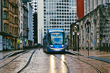 A blue tram, travels along tram tracks in a city setting. Modern and older buildings flank the street, which appears wet in Birmingham, UK.