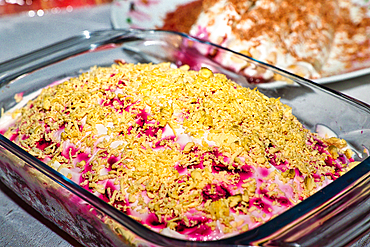 A glass dish contains a layered salad topped with shredded egg yolks. The salad layers are visible beneath, showing pink beetroot and white creamy elements. A blurred background shows another dish. Russian food.