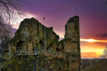 Ruins of a stone castle at sunset. The castle walls are partially collapsed, showing aged stonework. A flagpole stands within the ruins, and a lamppost is visible in the foreground. The sky displays vibrant orange and purple hues in Knaresborough, UK.