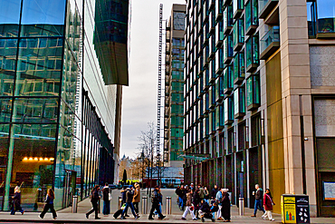 Modern city street scene with pedestrians walking between tall glass and stone buildings. A tall construction lift is visible in the background. The scene is brightly lit in London, UK.