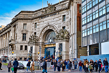 Waterloo Station's main entrance, a grand stone building with classical architecture, is shown with numerous people walking around the entrance. A modern building flanks the station to the right in London, UK.