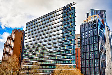 A CitySuites building, predominantly dark-glass and angular, stands prominently amongst other modern high-rises, including a bronze-colored building, under a partly cloudy sky in Manchester, UK.