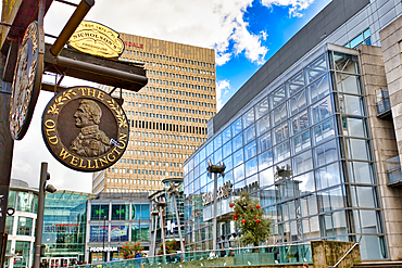 Urban scene featuring The Old Wellington pub sign, a modern glass building, and a high-rise building in the background. The vibrant colors and varied architectural styles create a dynamic composition in Manchester, UK.