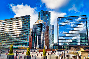 Modern high-rise buildings and a bridge in a city setting under a partly cloudy blue sky. Glass facades reflect the sky and clouds. Pedestrians are visible on a nearby walkway in Manchester, UK.