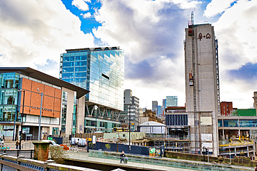 Urban cityscape featuring modern glass buildings, a taller concrete structure undergoing construction with scaffolding and an external lift, and a Harve Nichols department store. Partial view of a cityscape in the background in Manchester, UK.