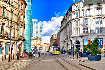 A sunny city street scene features a yellow tram traversing tram tracks, flanked by Victorian and modern buildings. Pedestrians populate the sidewalks, with businesses lining the street. The sky is bright blue with fluffy white clouds in Manchester, UK.